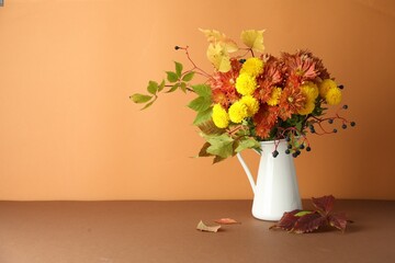 Autumn atmosphere. Vase with beautiful flowers, berries and leaves on brown table against orange background, space for text