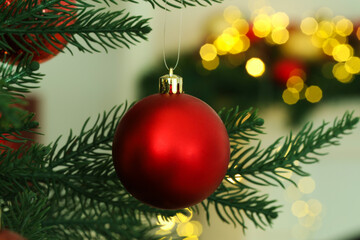 Red Christmas bauble hanging on fir tree branch against blurred lights, closeup. Bokeh effect