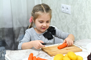 Little girl cuts carrot with knife on wooden board, fresh vegetables on table, kitchen background, cooking concept, child lifestyle activity. Girl slicing carrot with knife,