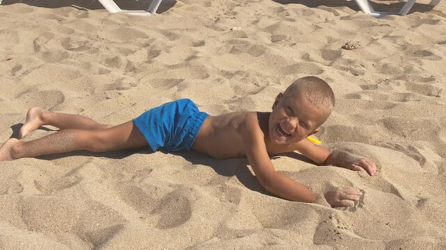 A young boy in blue swim shorts is lying prone on his stomach in the warm, golden sand and giving a cheeky grin at the camera. He is enjoying his time on a bright, sunny beach.