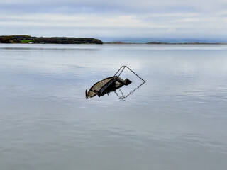 Remains of a sunken boat resting in calm reflective water beneath a soft overcast sky.