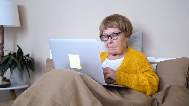 Senior woman in glasses typing on a laptop in bed, comfortably working or browsing online from home, staying connected with family, learning, or doing remote freelance tasks