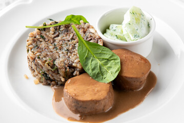 Gourmet dish with meat medallions, creamy sauce, and a side of groats or risotto with herbs and a separate bowl of cucumber salad