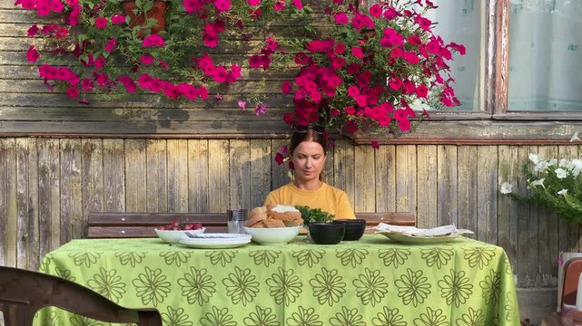 A woman in a yellow shirt sits at an outdoor table set with food, ready for a simple summer meal. She is framed by vibrant pink petunias against a rustic, weathered wooden dacha wall