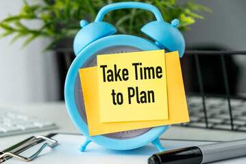 A blue alarm clock with a yellow note that says Take Time to Plan. The clock is placed on a desk with a keyboard and a pen