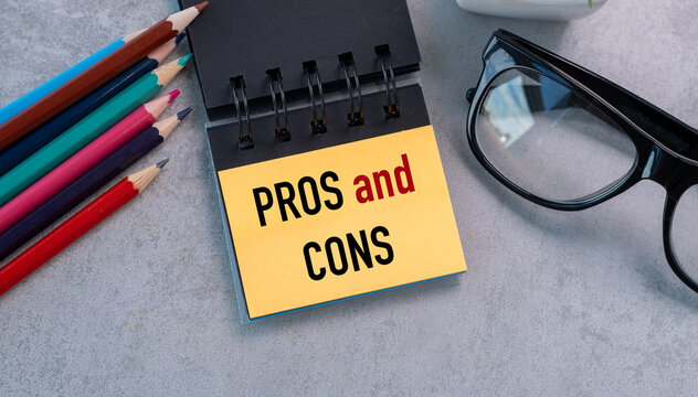 A notebook with the words Pros and Cons written on it. The notebook is on a table with a pencil, a pen, and a pair of glasses