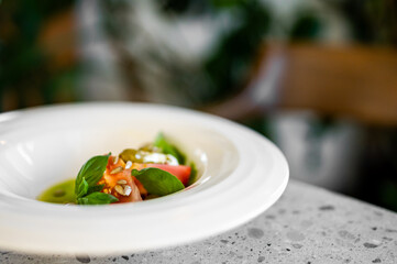 A vibrant, fresh vegetable salad with creamy white cheese, basil, and pumpkin seeds on a light-colored countertop. The meal is served in a white bowl at a restaurant