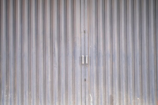 Industrial-style metal folding gate with vertical corrugated lines and central handles, commonly used for shopfronts or warehouse entrances.
