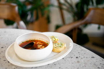 A bowl of rich seafood soup with mussels and shrimp is served with toasted bread on a modern countertop. The background is a soft-focus interior with plants