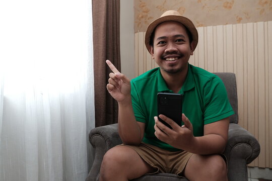 Happy man in green shirt and hat sitting on a chair, holding a smartphone in one hand and pointing upward with a cheerful smile.