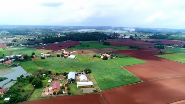 Aerial view of a sprawling rural landscape with a patchwork of green fields, reddish soil, scattered homes, and distant hills
