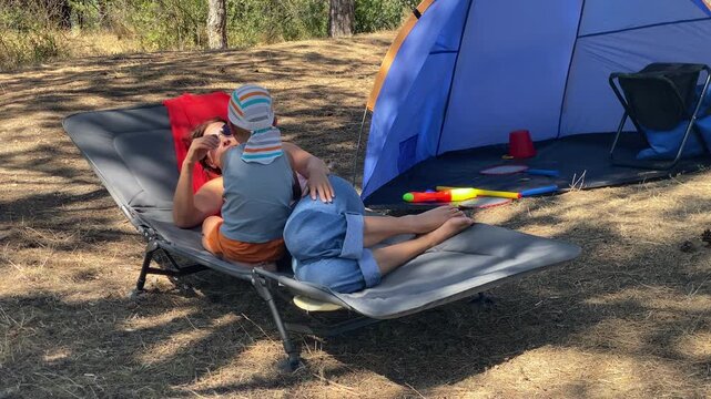 A mother smiles while lovingly hugging her young son as they lie together on a camping cot. They are resting by their tent in a sunny, forested area.