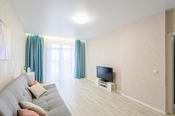 A bright living room with a gray sofa, teal curtains, and a TV. The room has light walls and wooden floors. Natural light floods the space