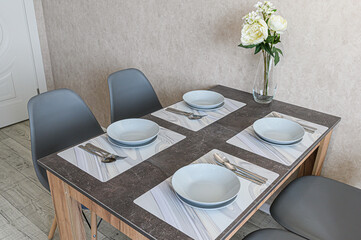 A modern dining table is set for four with bowls, silverware, and placemats. A vase of white flowers adds a touch of elegance