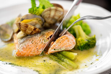 A close-up of a fork and knife cutting into a perfectly cooked salmon fillet on a white plate. The dish is served with clams, green asparagus, and broccoli in a light sauce