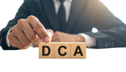 A suited person arranges wooden blocks spelling "DCA", symbolizing financial concept