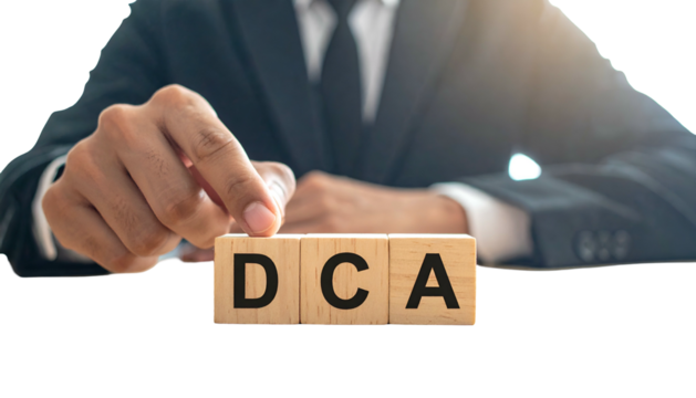A suited person arranges wooden blocks spelling "DCA", symbolizing financial concept