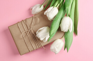 Gift box wrapped in brown paper with twine, adorned with fresh white tulips, resting on a soft pink background, creating a vibrant and inviting atmosphere for celebrations