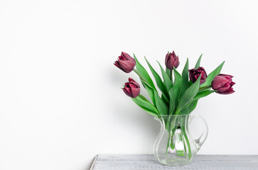 Purple tulips arranged in a clear glass vase on a rustic wooden table against a soft white background, creating a serene and elegant floral display for home decor