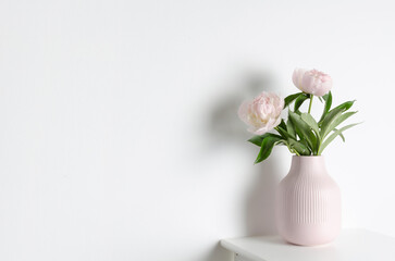 Elegant pink vase with fresh peony flowers sits on a minimalist white table against a clean white wall, creating a serene and stylish home decor atmosphere