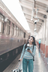 Obraz premium Asian woman with backpack looking forward while standing at train station, holding luggage, waiting for train to depart at busy station, excited about the journey.