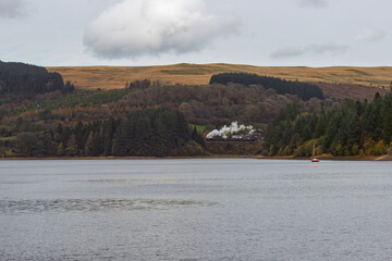 A vintage steam locomotive puffs smoke along the Brecon Mountain Railway, winding around the Pontsticill Reservoir amidst the autumnal beauty of Bannau Brycheiniog (Brecon Beacons National Park) Wales