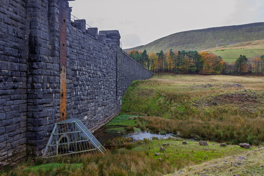The internal stone wall of the decommissioned Upper Neuadd Reservoir with its restricted intake tunnel and slow flowing Taf Fechan river in Bannau Brycheiniog (Brecon Beacons), Powys, Wales