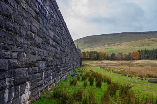 A historic dam stone wall of the Grade II listed Upper Neuadd Reservoir stretches into the distance with autumn trees lining the Taf Fechan river basin in Bannau Brycheiniog (Brecon Beacons), Powys