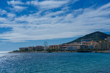 Fototapeta premium Coastal cityscape with colorful buildings, Ferris wheel, and sea under bright sky and scattered clouds.