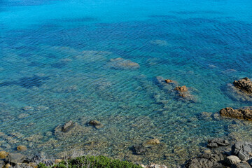 Shallow turquoise water along a rocky coast with visible underwater stones and sea plants. Clear Mediterranean landscape viewed from above.