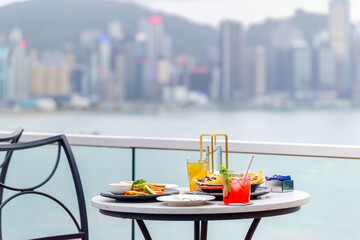 Outdoor dining table overlooking Hong Kong skyline with colorful cocktails and appetizers on sunny day. Urban dining experience, cityscape backdrop, relaxed atmosphere.