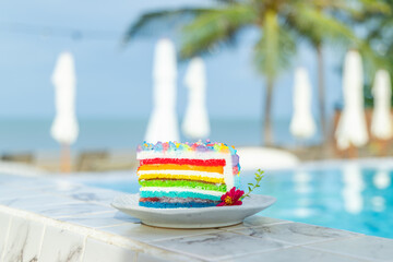 Colorful rainbow cake by pool at tropical resort, vibrant dessert with ocean view, perfect for...