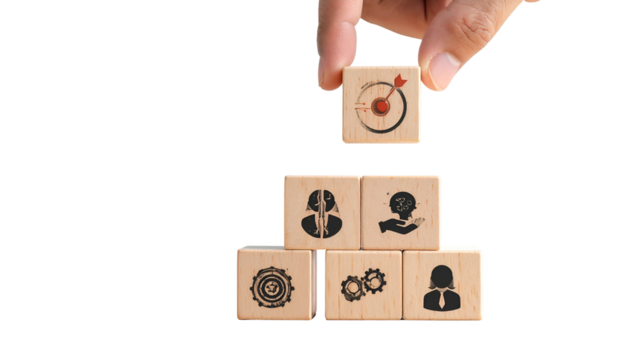 Hand placing a wooden block with target icon atop a pyramid of blocks with business icons