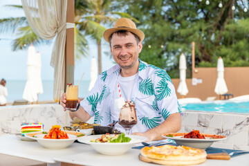 Smiling man wearing tropical shirt and hat enjoying drinks and diverse food at beachside restaurant. Tropical vacation, relaxation, culinary delight with beach and palm trees in background.