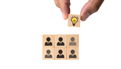 Hand placing a lightbulb icon block above blocks with human figures, symbolizing teamwork