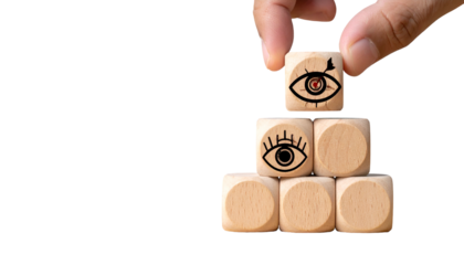 Hand adding wooden eye cube to pyramid of similar cubes on black, emphasizing vision & perspective
