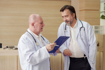 Male doctors reviewing patient medical file together during hospital team meeting at reception desk discussing diagnosis plan and coordinating urgent care for improved treatment and patient recovery