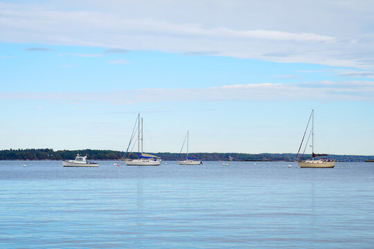 Three sailboats and a small motorboat float in the calm waters of Portland Harbor, Maine, against a clear blue sky and distant tree-lined shoreline, symbolizing peace and maritime leisure.