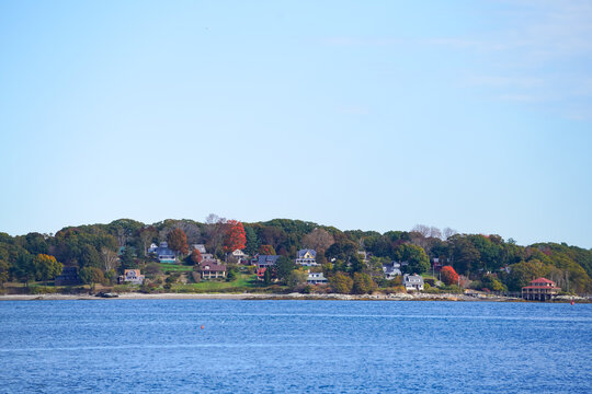 A scenic coastal neighborhood viewed from across calm blue water, featuring colorful autumn trees and waterfront homes beneath a bright open sky.