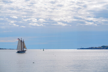 A schooner sails across calm coastal waters beneath bright clouds, its sails glowing in soft sunlight as it moves toward the horizon on a peaceful day at sea.