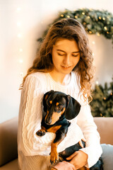 beautiful curly girl in a white sweater with dark hair sits on an armchair and holds a black dachshund in her arms. happy New Year's portrait on the background of a Christmas tree and a white window
