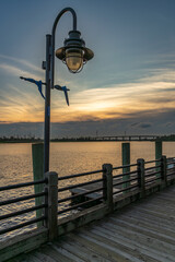 Fototapeta premium A peaceful sunset illuminates the Wilmington, North Carolina Riverwalk, where a vintage lamppost stands over a wooden boardwalk beside the calm Cape Fear River.