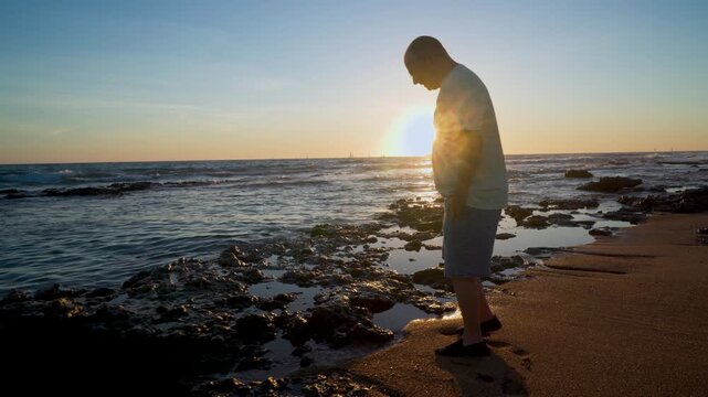 Hombre mayor mirando la orilla del mar al atardecer con el sol detr&aacute;s de el.