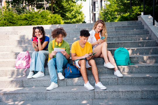 Young group of friends sitting on steps outdoors, enjoying social media, studying, and bonding during the sunny day - Powered by Adobe