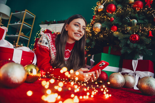 Joyful Christmas moment a smiling woman in a red sweater enjoys a cozy home scene with a decorated tree and gifts while using a smartphone - Powered by Adobe