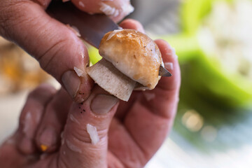 A close-up view captures a person's hands carefully peeling the skin off a small mushroom with a knife, set against a soft, out-of-focus green background.