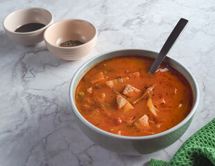 A bowl of soup with a spoon in it sits on a marble countertop. The bowl is filled with a red and green soup, and there are two bowls with spices. Concept of comfort and warmth