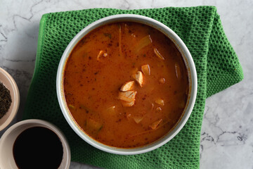 A table top view of bowl of soup with a green cloth. bowl is filled with a red soup and has a few pieces of tofu in it. Bowl of soy sauce on the side