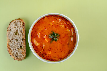 A bowl of soup sits on a table next to a piece of bread. The soup is red and has a lot of vegetables in it. he soup is sprinkled with parsley
