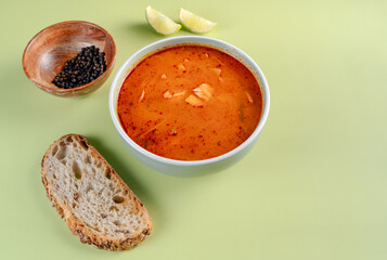 A bowl of soup sits on a table next to a piece of bread. The soup is orange and has a spiced flavor. The bread is to the left of the soup and he is a simple, plain loaf